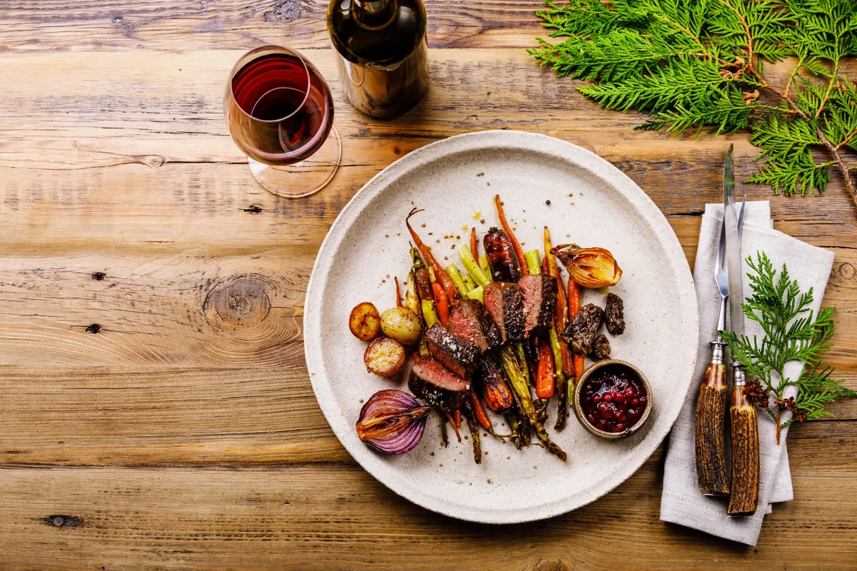 Wildgericht mit Gemüse, Preiselbeeren und Rotwein auf rustikalem Holztisch